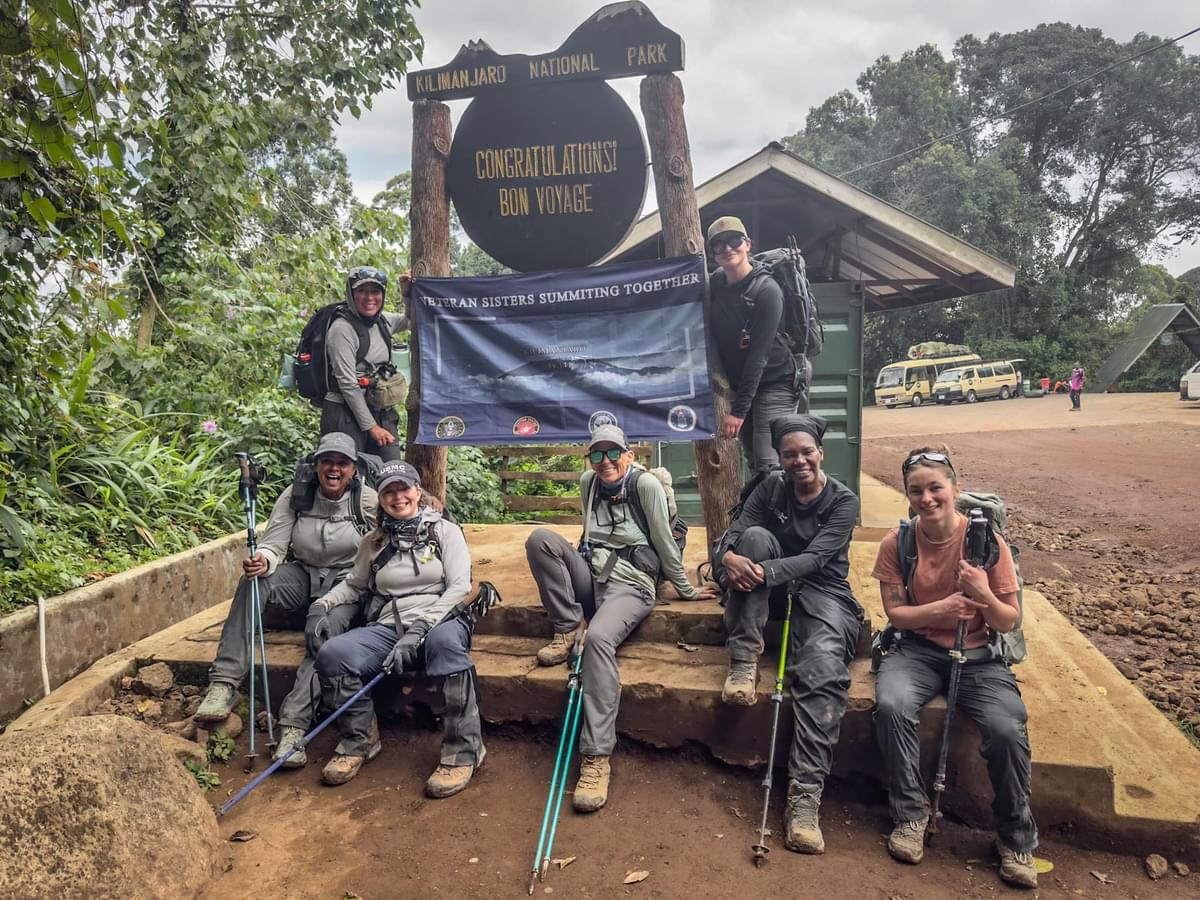 April Harris with her tribe at the Kilimanjaro National Park 'Congratulations · Bon Voyage' gate, holding a 'Veteran Sisters Summiting Together' banner.